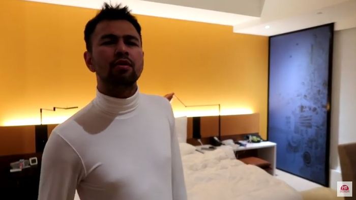 Raffi Ahmad berdiri di depan sebuah tempat tidur yang ada di dalam kamar hotelnya.