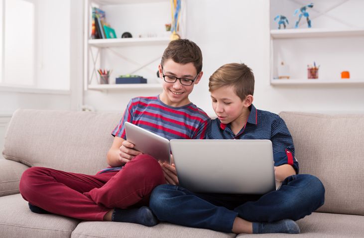 Two male kids with gadgets. Sharing funny content on laptop and digital tablet on sofa at home. Boy friendship and communication concept