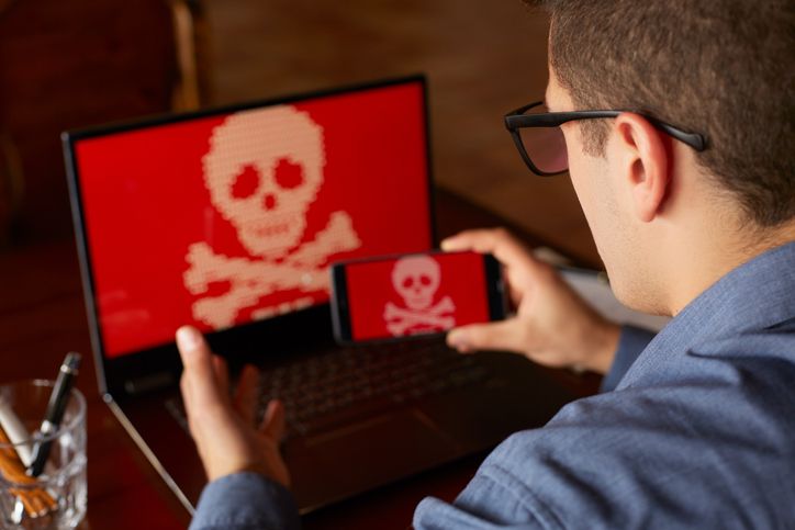 Man sits near laptop with phone blocked and encrypted by ransomware spyware asking for money. Laptop and smartphone infected by virus. Scary red skull crossbones on screen. Cyber security theme.