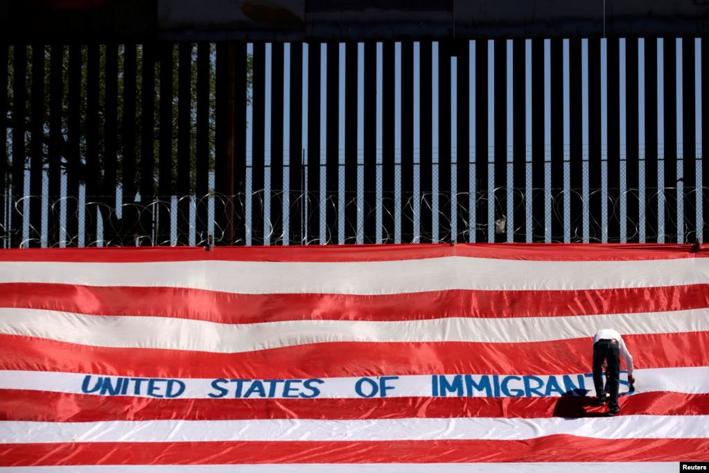 Roberto Marquez, yang dikenal sebagai Roberz, menulis pada sebuah bendera AS berukuran besar sebagai bagian dari unjuk rasa yang disebut ‘United States of Immigrants’ untuk menuntut penghormatan kepada kaum migran, dekat tembok perbatasan di El Paso, Texas, sebagaimana tampak dalam gambar dari Ciuda