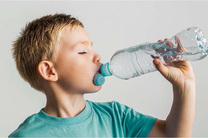 Seorang anak yang gembira sedang minum air putih setelah bermain di luar, menunjukkan pentingnya minum untuk menjaga tubuhnya tetap terhidrasi