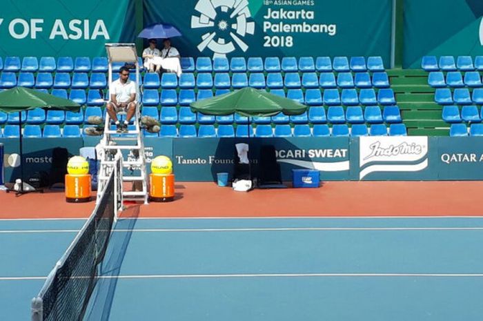 Dua penonton cabang olahraga tenis berbagi payung di lapangan tenis kompleks Jakabaring Sport City, Palembang, pada hari Rabu (22/8/2018).