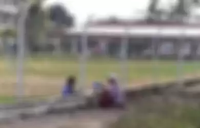 Seorang nenek duduk bersama cucunya di bawah pohon untuk makan siang, namun mereka dipisahkan oleh pagar besi sekolah